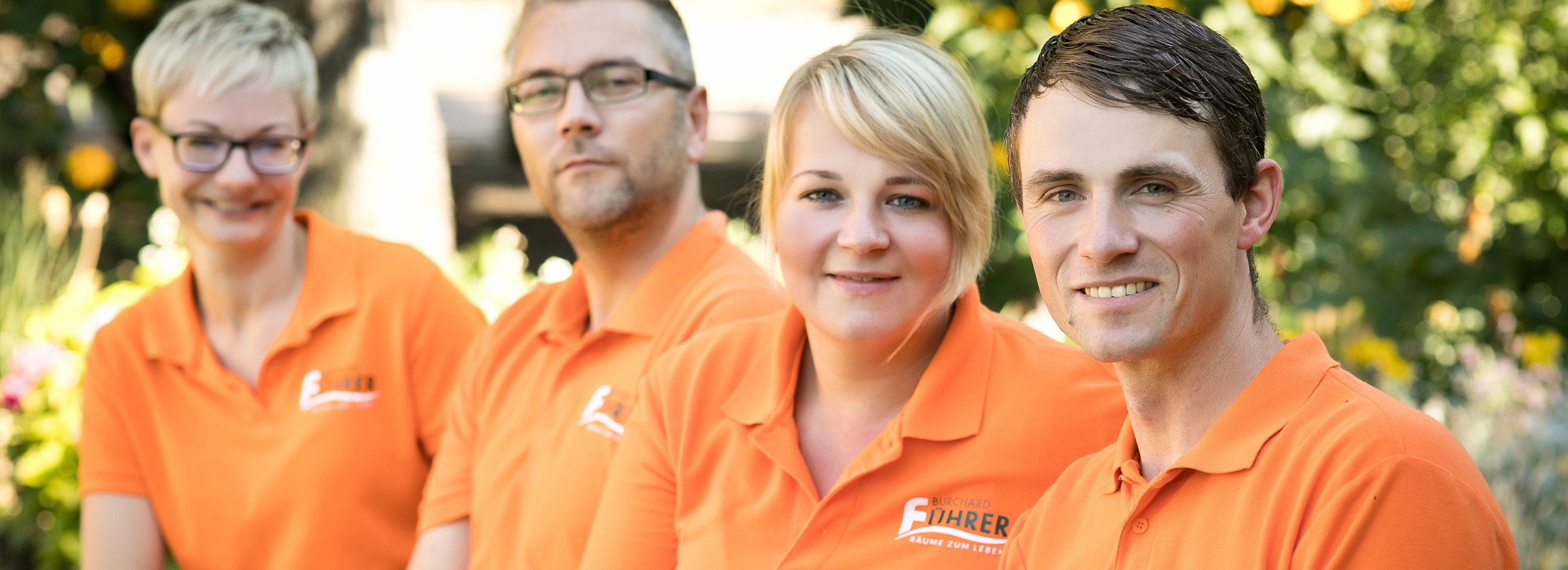 Titelbild Informationen 4 Mitarbeiter in orangenen Polo sitzen auf einer Bank und schauen freundlich in die Kamera